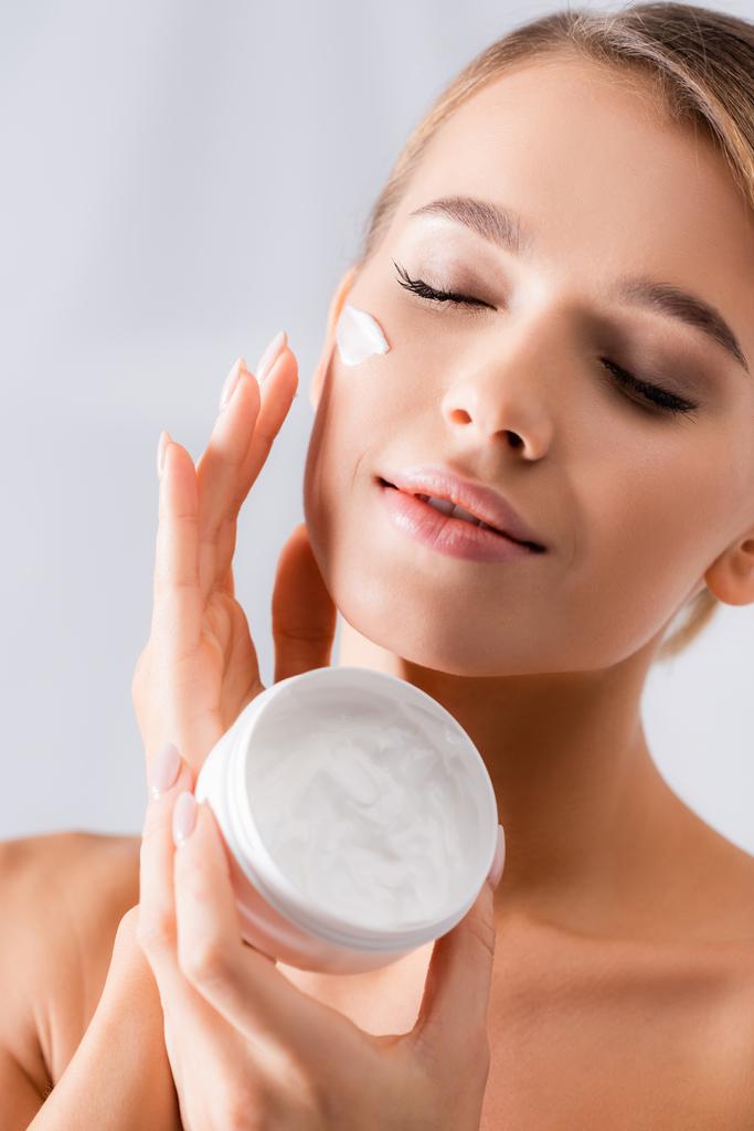 woman with bare shoulders holding jar and applying face cream on white - Photo, Image