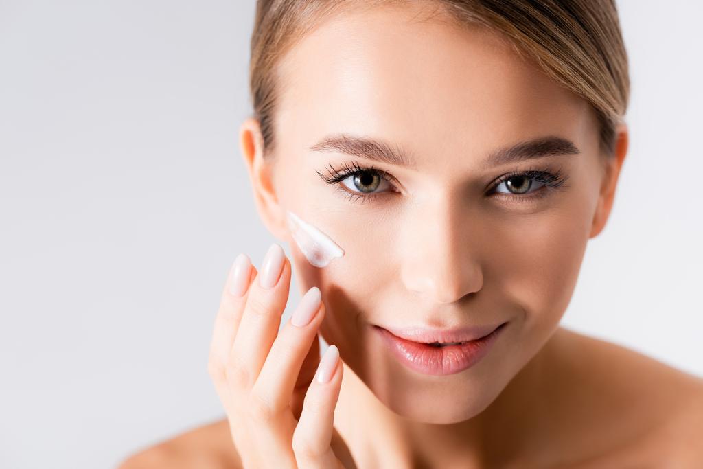 radostná mladá žena nanášení hydratační krém na obličej izolované na bílém - Fotografie, Obrázek
