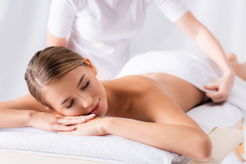 massagista desfocado ajustando toalha no cliente com os olhos fechados deitado na mesa de massagem - Foto, Imagem