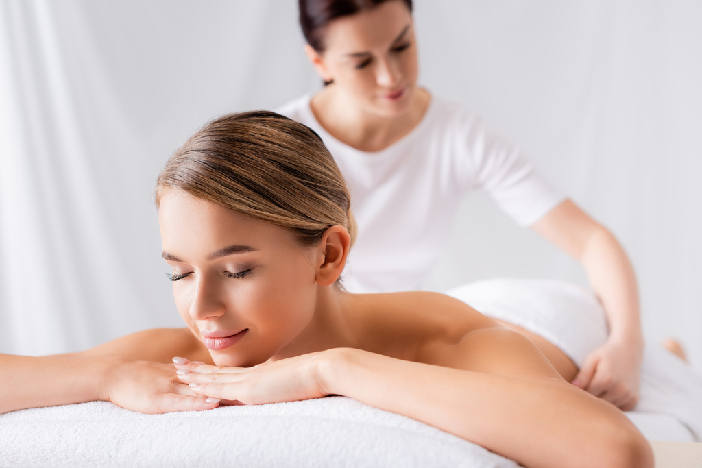 jovem mulher deitada na mesa de massagem perto de massagista no fundo borrado - Foto, Imagem