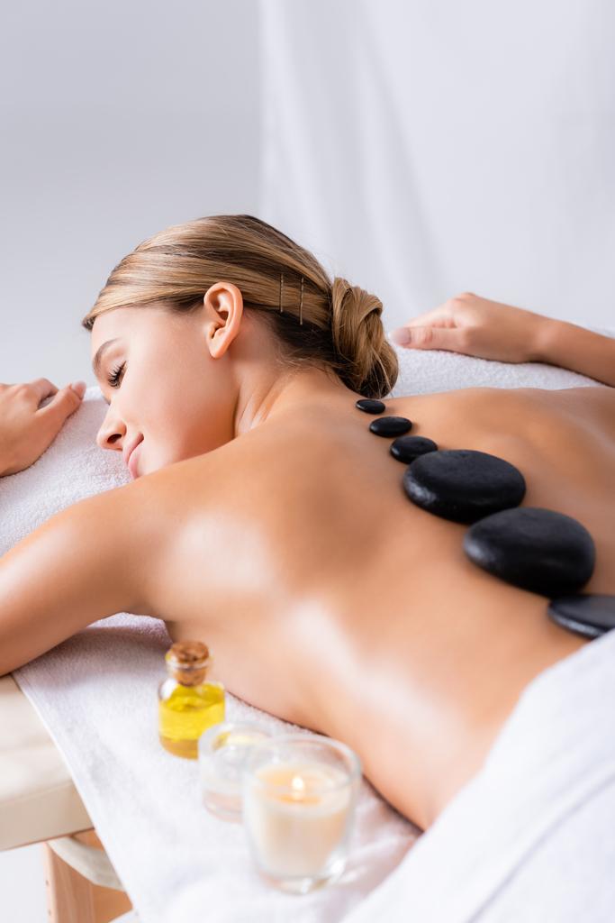 junge Frau mit geschlossenen Augen bekommt Hot-Stone-Massage bei brennender Kerze im Wellness-Salon  - Foto, Bild