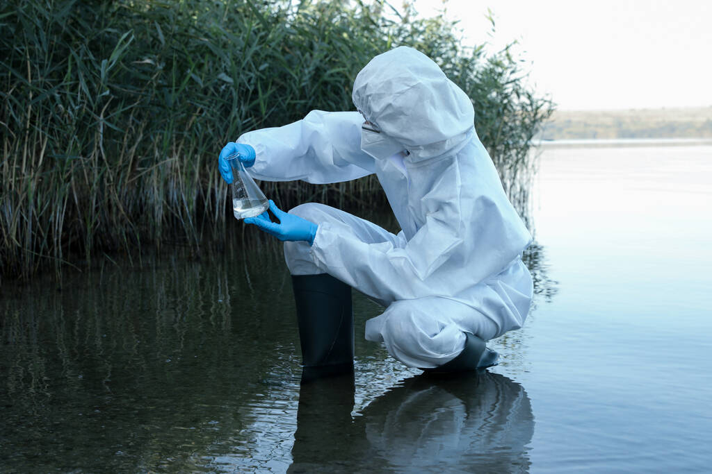 Naukowiec w chemicznym kombinezonie ochronnym z kolbą stożkową pobierający próbki z rzeki do analizy - Zdjęcie, obraz