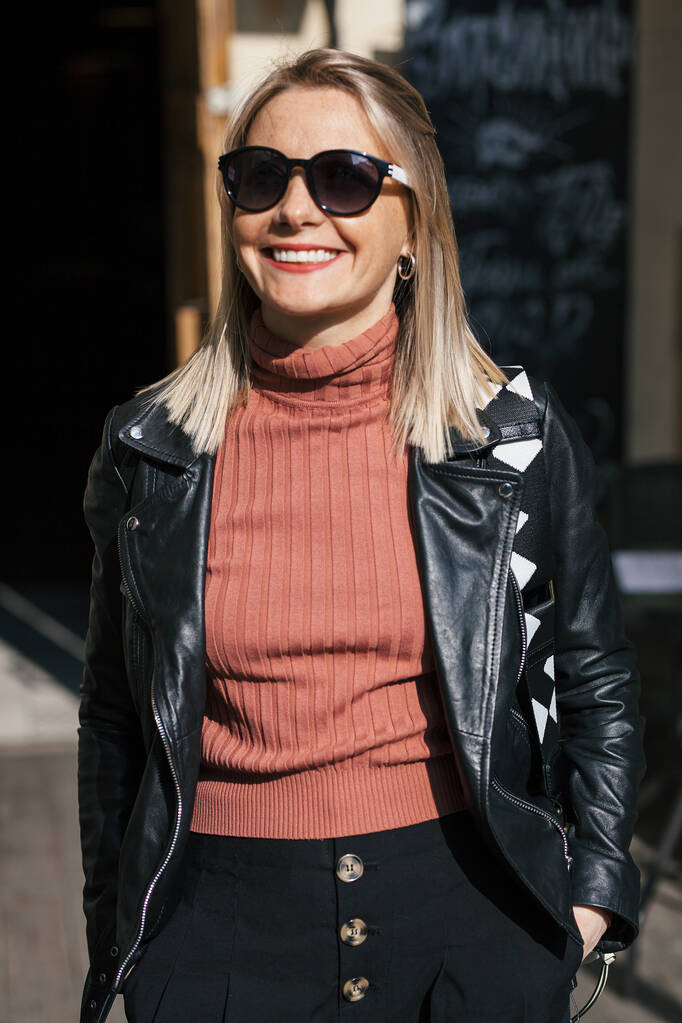nuori kaunis hymyilevä tyylikäs blondi nainen aurinkolasit, mustat housut ja nahkatakki poseeraa rakennusten don kaupunkiympäristössä - Valokuva, kuva