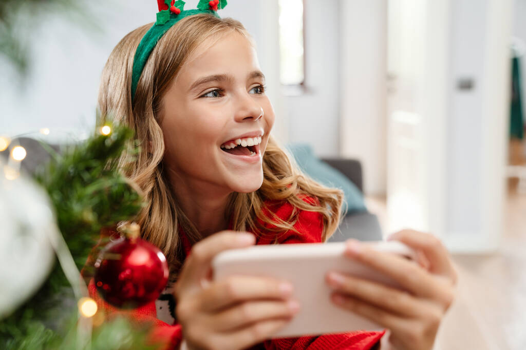 Menina caucasiana feliz usando chifres de veado de brinquedo jogando videogame no celular em casa - Foto, Imagem