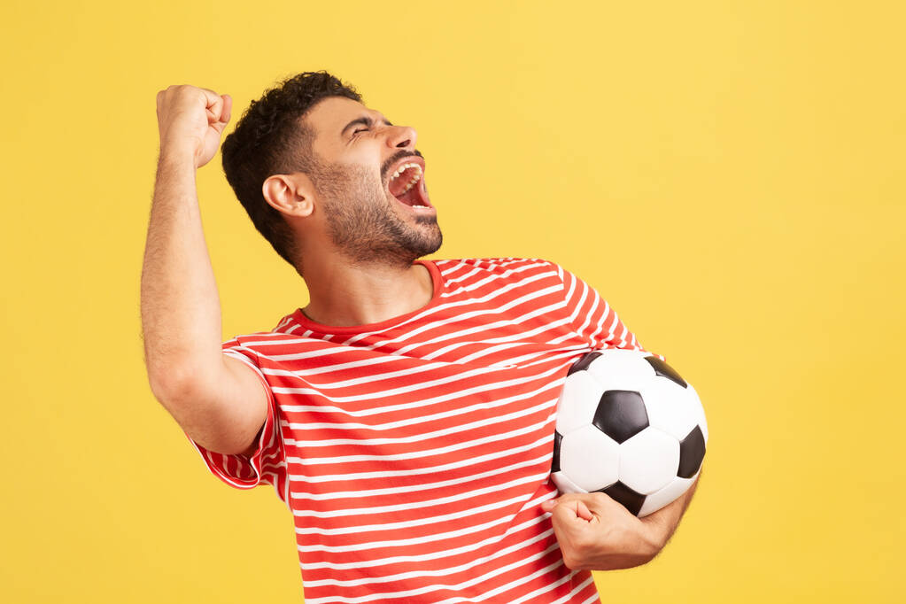 ストライプの赤いTシャツを着た非常に肯定的な満足した男は、チャンピオンシップで好きなサッカーチームの勝利を祝う、サッカーボールを保持叫んでいます。黄色の背景に隔離された屋内スタジオショット - 写真・画像
