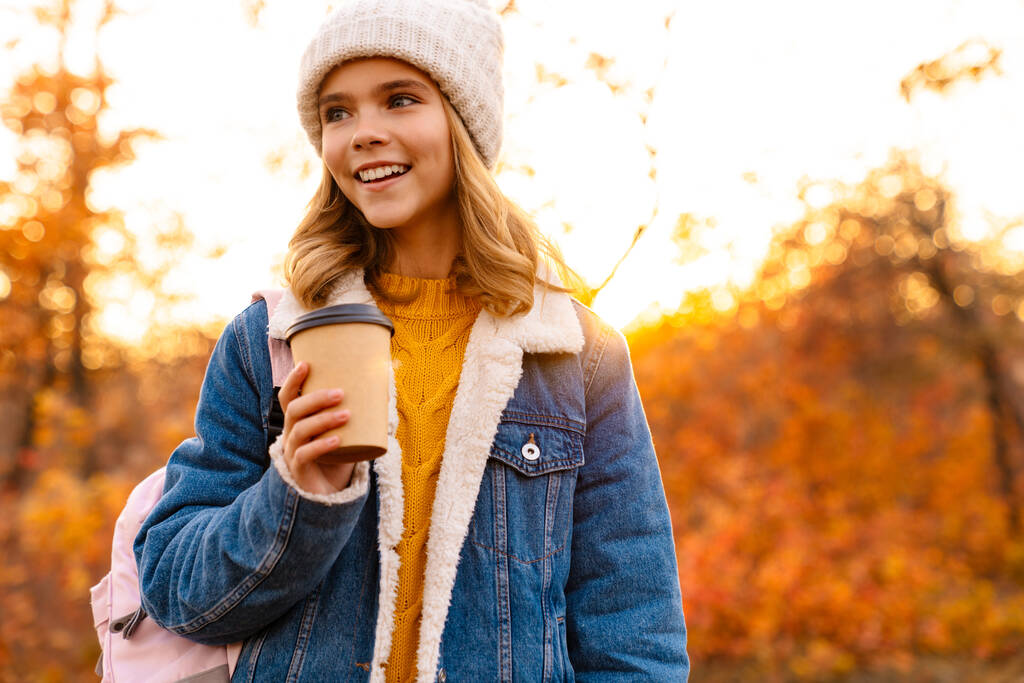バックパックを持って公園を歩いて秋の服を着て笑顔の女の子,コーヒーカップを保持 - 写真・画像