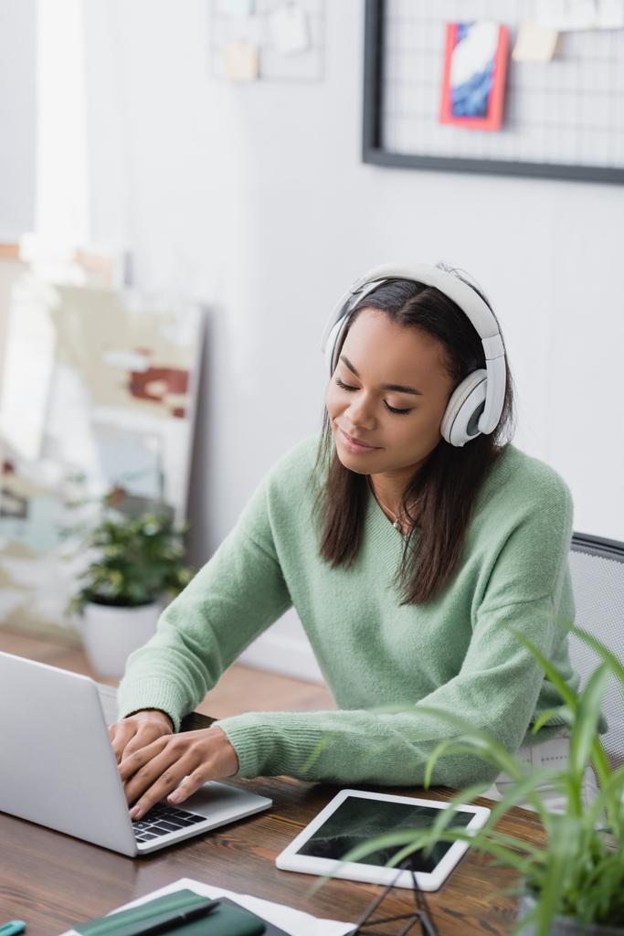 positive african american interior designer in headphones typing on laptop at home, blurred foreground - Photo, Image