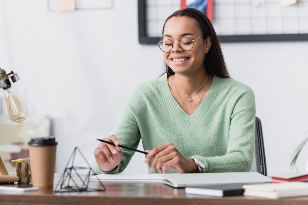 heureux architecte afro-américain dans les lunettes tenant crayon tout en étant assis au bureau sur le premier plan flou - Photo, image