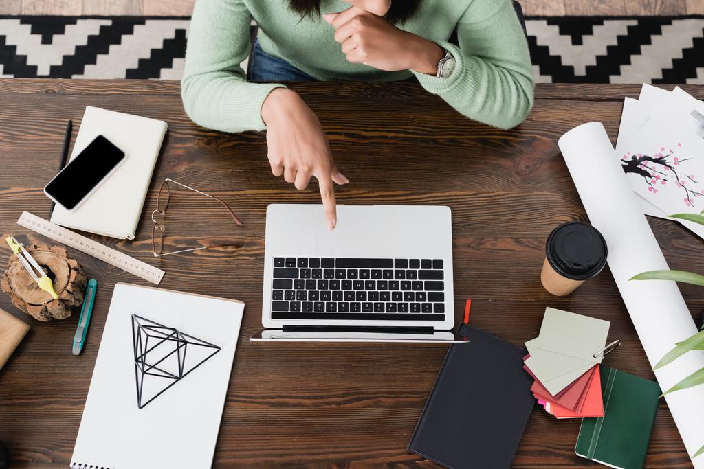 вид сверху на африканского американского дизайнера интерьера, указывающий на ноутбук рядом с моделью пирамиды, смартфон и бумажный стаканчик на столе - Фото, изображение