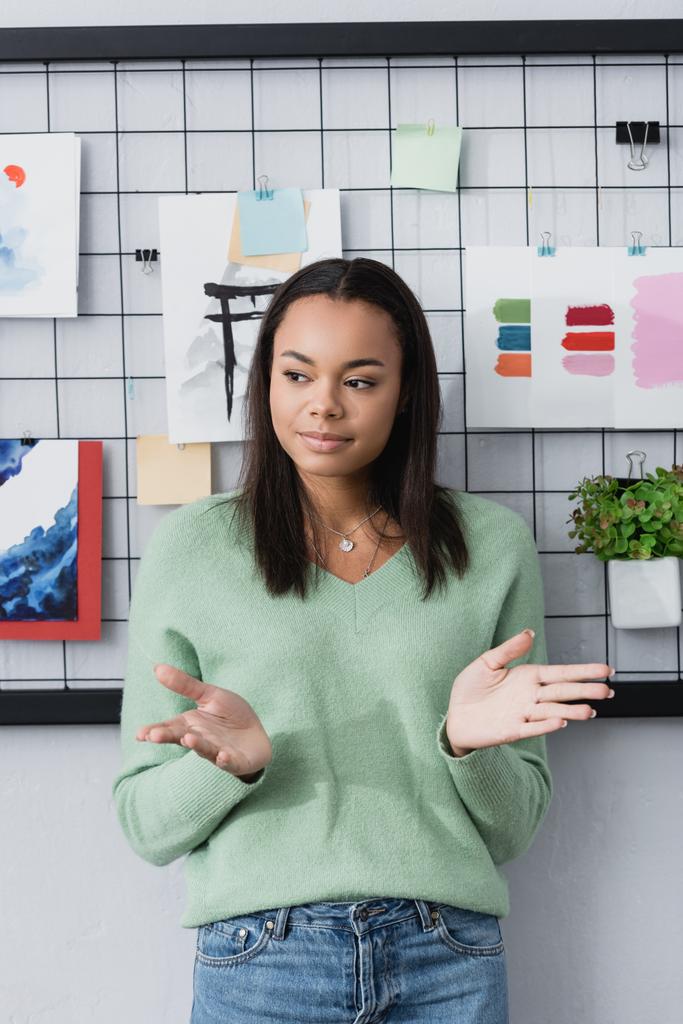mladá africká americká návrhářka interiérů gestikuluje, zatímco stojí u obrazů na stěně - Fotografie, Obrázek