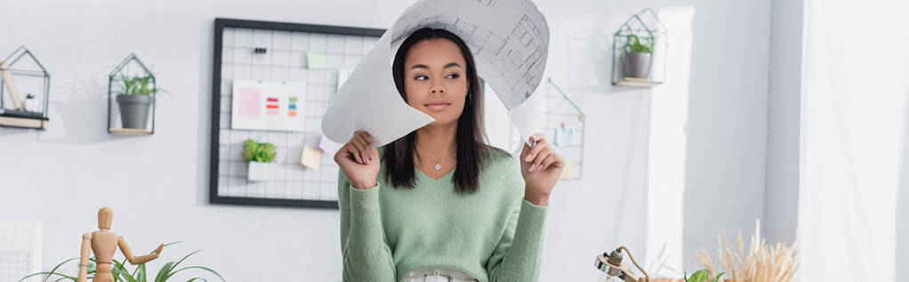 joven arquitecto afroamericano cubriendo la cabeza con un plano mientras mira hacia el hogar, pancarta - Foto, imagen