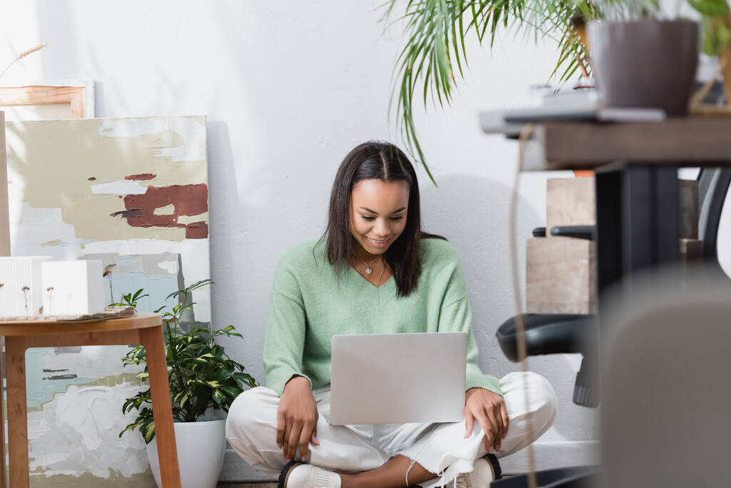 feliz arquiteto afro-americano sorrindo enquanto sentado no chão com laptop em estúdio em casa em primeiro plano borrado - Foto, Imagem