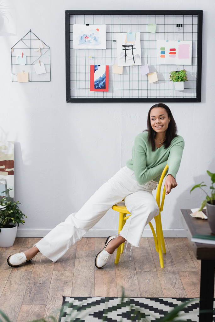 sorridente designer de interiores afro-americano olhando para longe enquanto sentado na cadeira em estúdio em casa, foreground borrado - Foto, Imagem