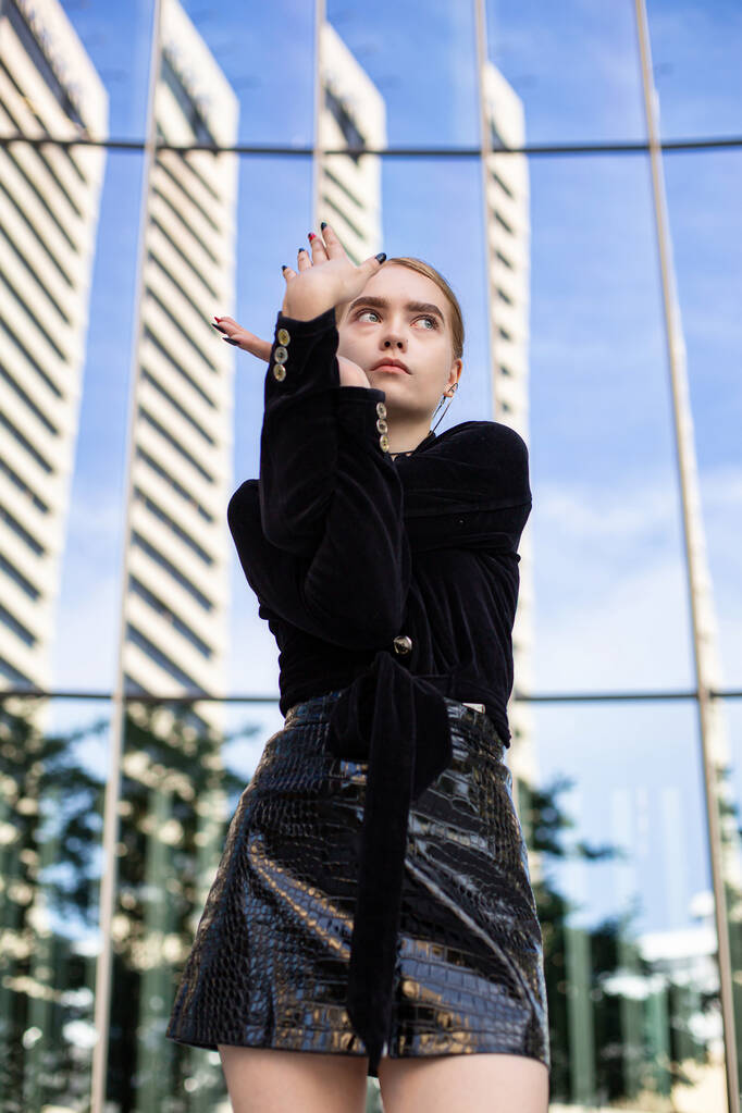 Молодая женщина с светлыми волосами стоит с рукой рядом с лицом в городе - Фото, изображение