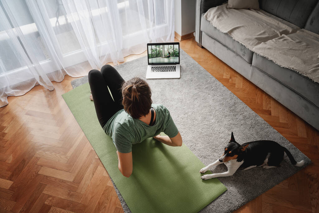 Весёлая красивая женщина практикующая йогу дома онлайн на ковре и йоге мат со своей собакой рядом, спорт и психическое здоровье - Фото, изображение