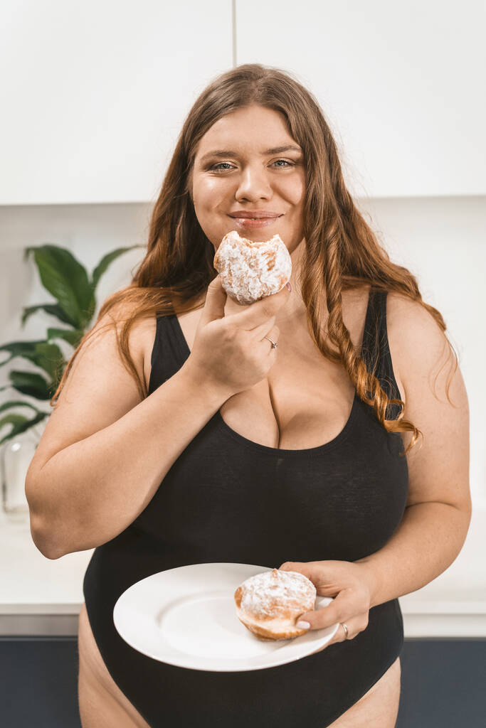 Zadowolona młoda gruba kobieta jedząca tort uśmiechnięta przed kamerą w czarnych strojach kąpielowych. Piękna pulchna młoda kobieta jedząca niezdrowe jedzenie. Fat girl jedzenie ciasto stojące na nowoczesnej kuchni - Zdjęcie, obraz