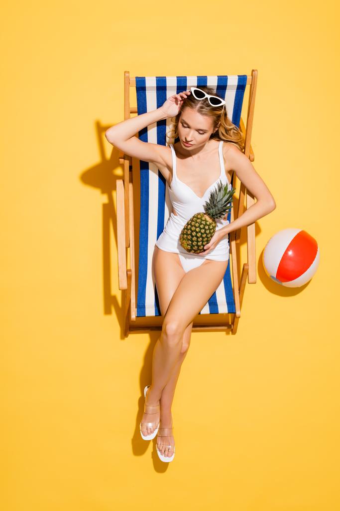 vue de dessus de la femme en maillot de bain ajustant les lunettes de soleil tout en étant assis dans la chaise longue avec ananas près ballon gonflable sur jaune - Photo, image