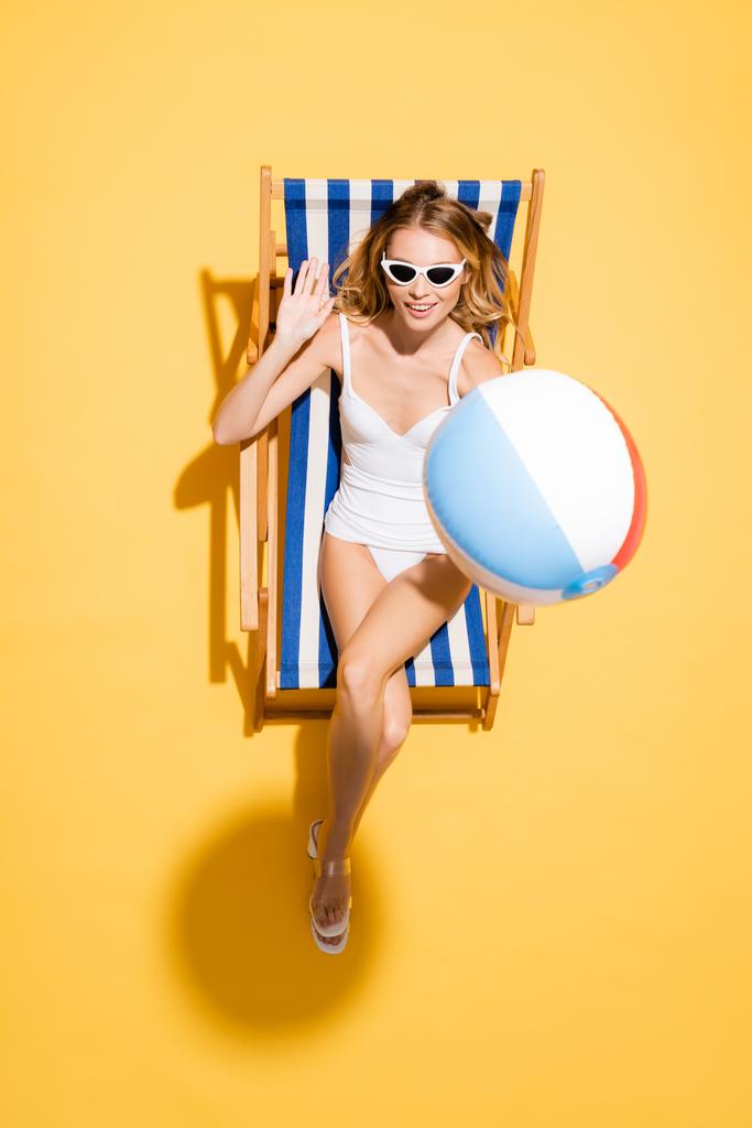 vrchní pohled na mladou ženu v slunečních brýlích a plavkách mávání rukou při relaxaci v lehátku s nafukovací koulí na žluté - Fotografie, Obrázek