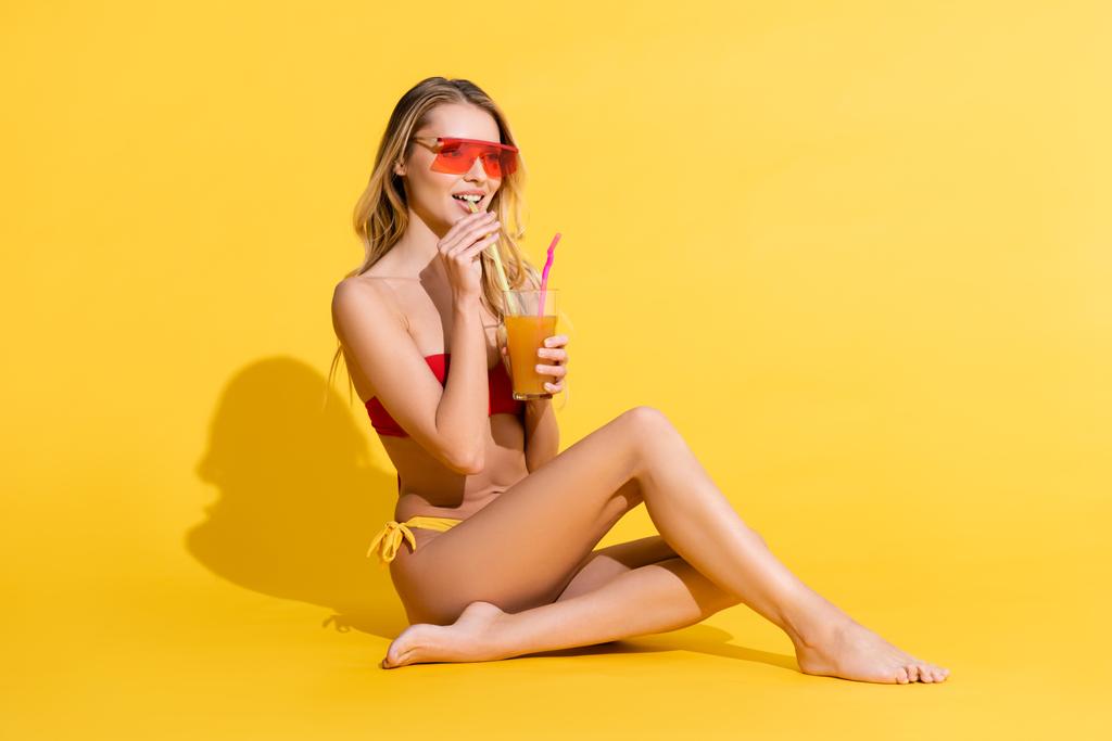 Улыбающаяся босиком женщина в купальнике смотрит в сторону и пьет свежий коктейль на желтом - Фото, изображение