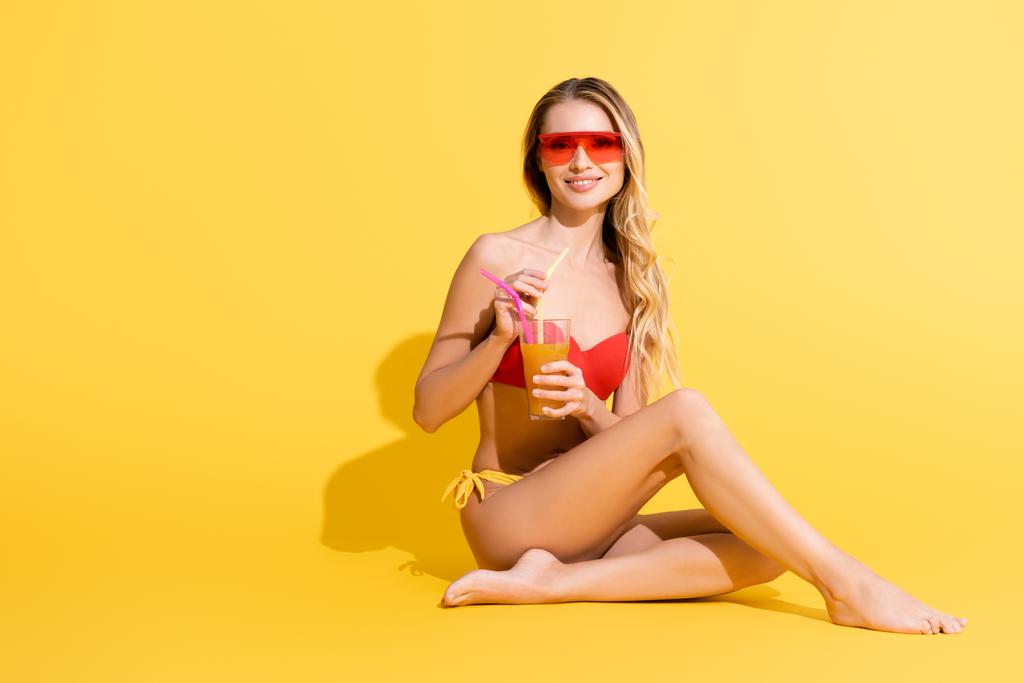 裸足の女性でサングラスをかけながらカメラで微笑む黄色のカクテルを持ち - 写真・画像