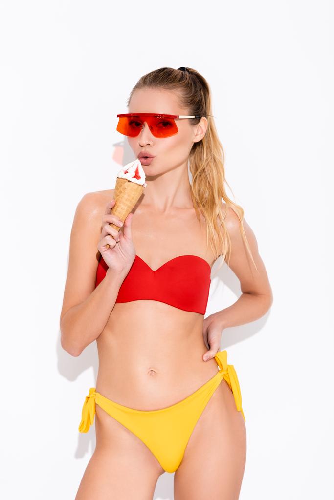 giovane donna in costume da bagno e occhiali da sole che tiene il gelato in cono waffle su bianco - Foto, immagini