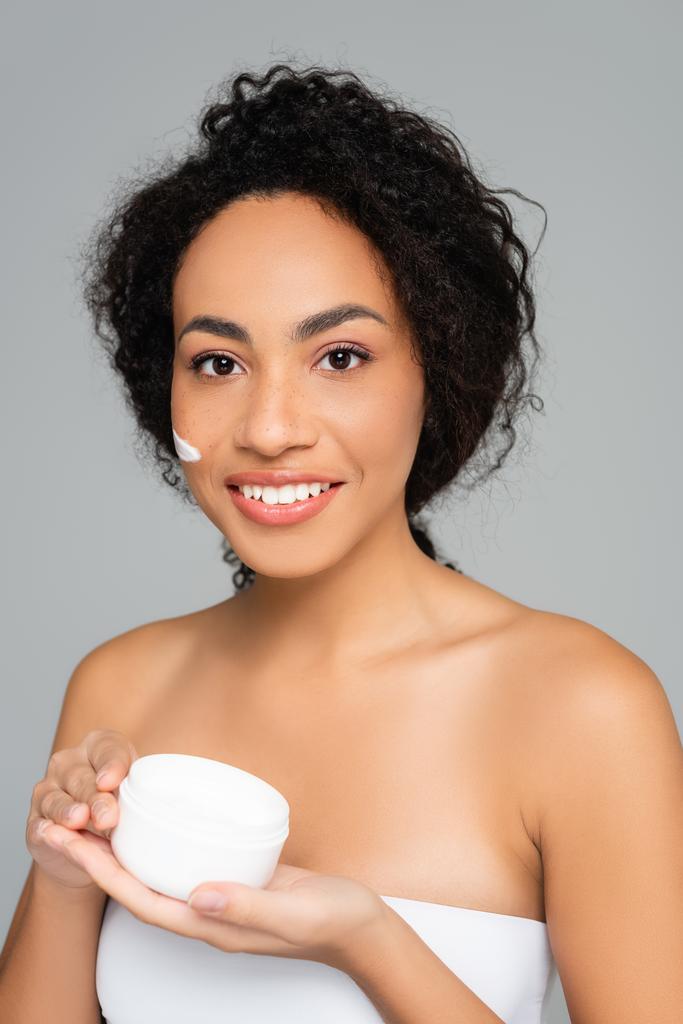 Afroamerikanerin lächelt, während sie Glas mit Kosmetikcreme auf grau isoliert hält  - Foto, Bild