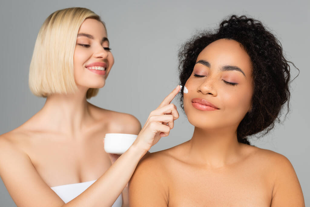 Afrikai amerikai nő áll közel szőke nő alkalmazó kozmetikai krém elmosódott háttér elszigetelt szürke  - Fotó, kép
