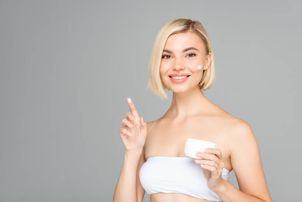 Mujer sonriente sosteniendo crema cosmética en el dedo aislado en gris  - Foto, Imagen