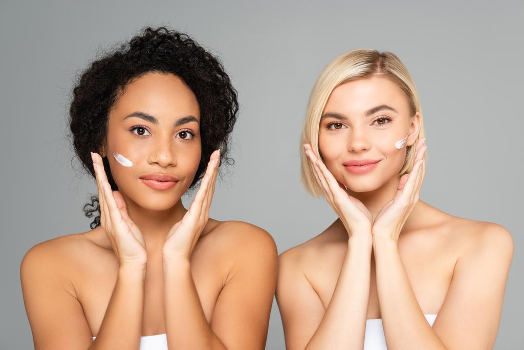 Mujeres multiétnicas con crema cosmética en las caras mirando a la cámara aislada en gris  - Foto, Imagen