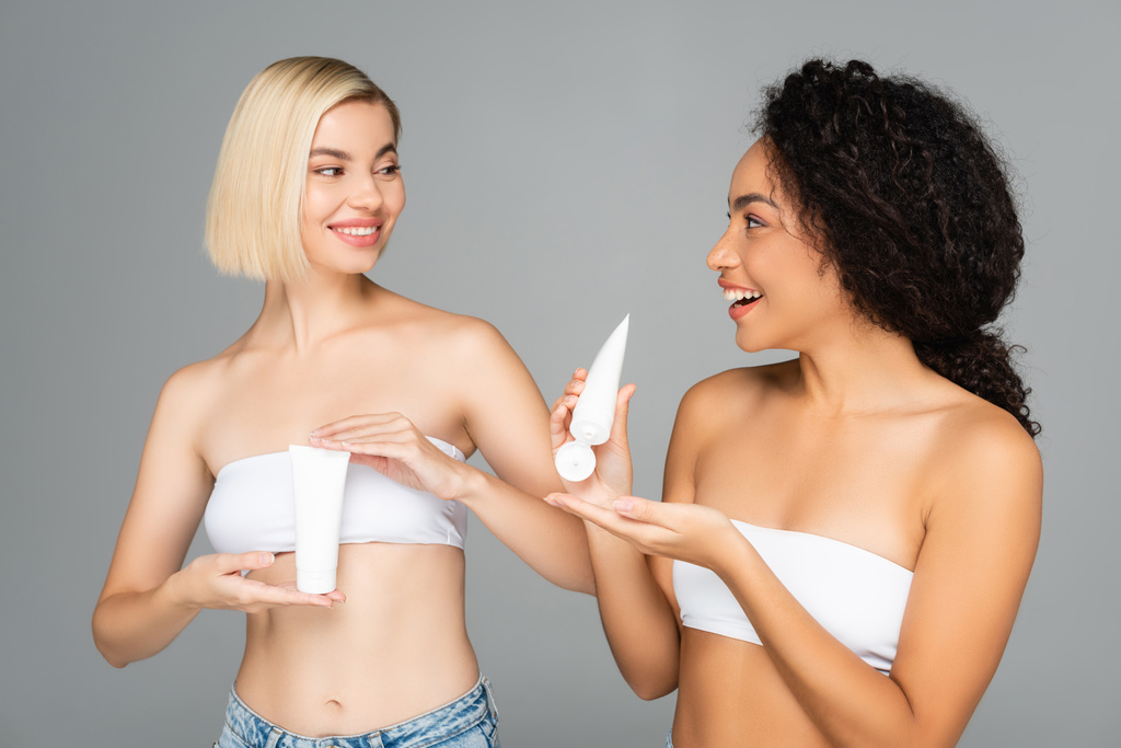 Mosolygó többnemzetiségű nők, akik szürke alapon izolált kozmetikai testápoló csöveket tartanak  - Fotó, kép