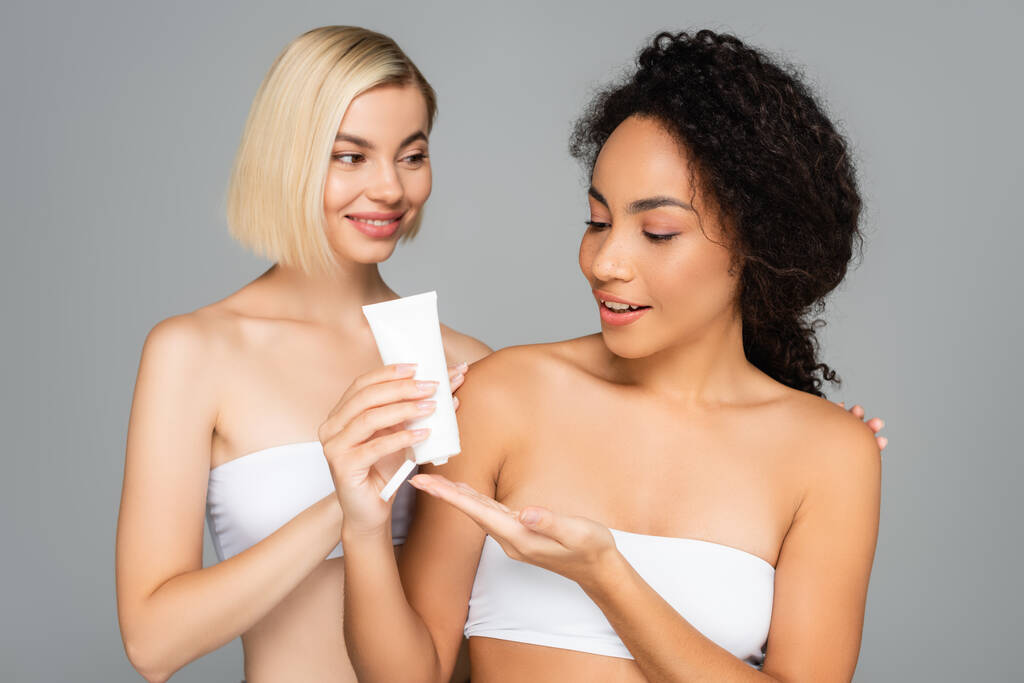 Femme blonde étreignant femme afro-américaine avec tube de crème cosmétique isolé sur gris  - Photo, image