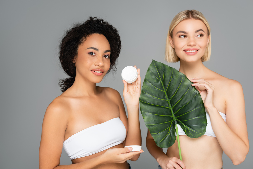 Улыбающиеся мультиэтнические женщины, держащие косметический крем и тематический лист, изолированный на сером  - Фото, изображение