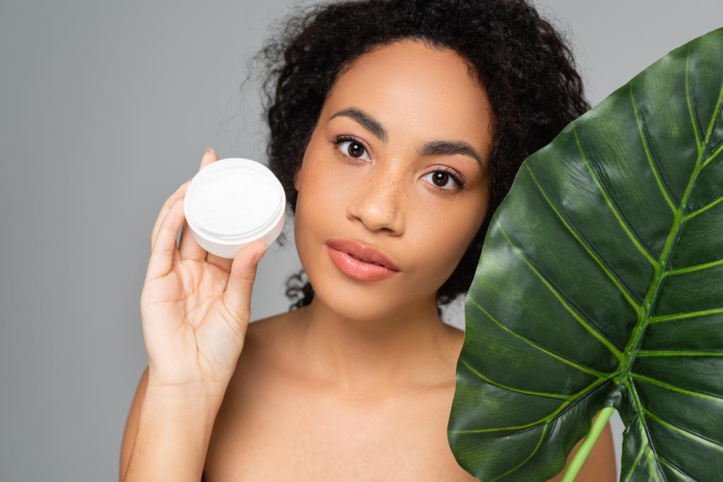 Afroamerikanerin mit perfekter Haut hält tropisches Blatt und kosmetische Creme isoliert auf grau  - Foto, Bild