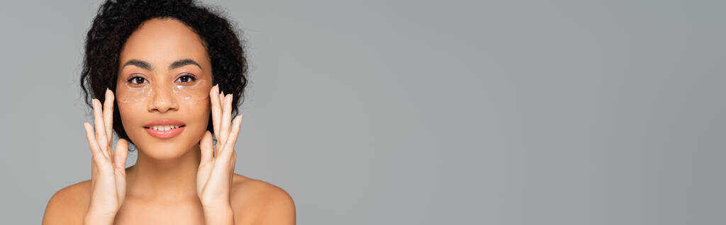 Mujer afroamericana con hombros desnudos aplicando parches oculares aislados en gris, pancarta  - Foto, imagen
