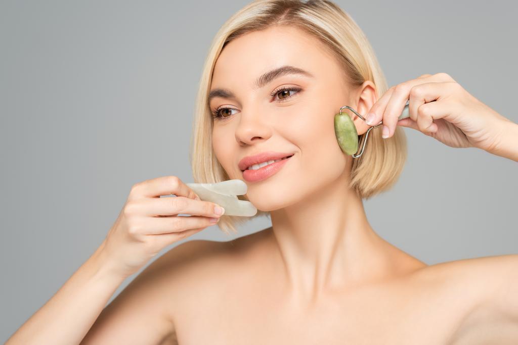 Blonde woman with clean skin massaging face with gua sha and jade roller isolated on grey  - Photo, Image