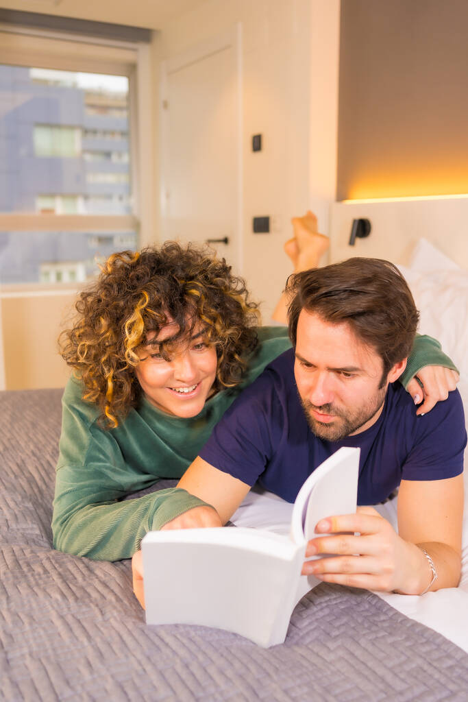 Estilo de vida, una joven pareja caucásica con miradas conocedoras, en pijama en la cama leyendo un bonito libro abrazando, foto vertical - Foto, imagen