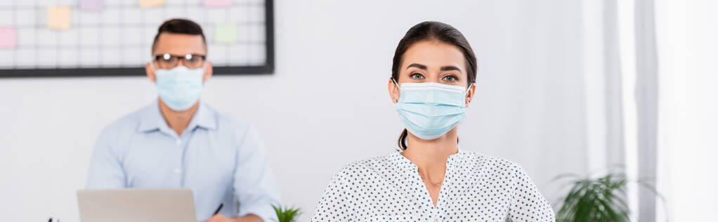 podnikatelka v lékařské masce při pohledu do kamery v blízkosti podnikatele na rozmazaném pozadí, banner - Fotografie, Obrázek