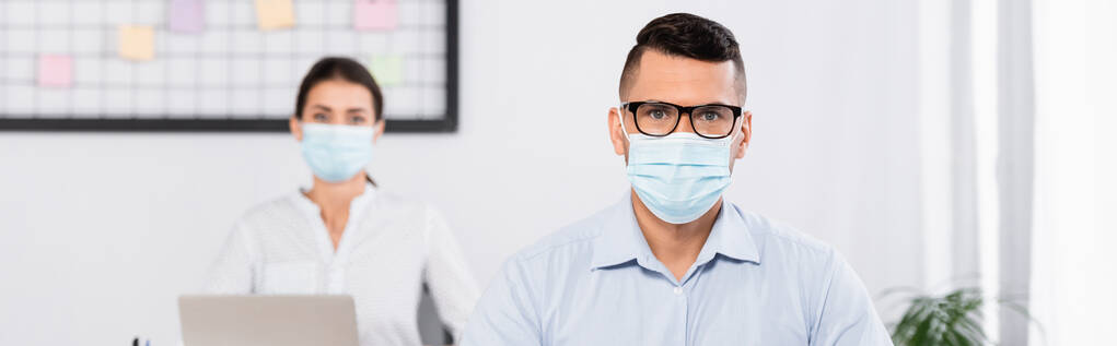 背景のぼやけたビジネスウーマンの近くのカメラを見ている医療用マスクや眼鏡のビジネスマン - 写真・画像