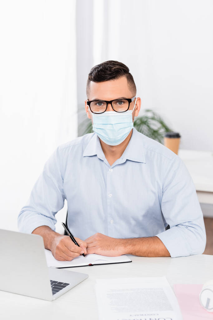 бизнесмен в медицинской маске и очках смотрит в камеру, держа ручку возле ноутбука и ноутбука на столе - Фото, изображение