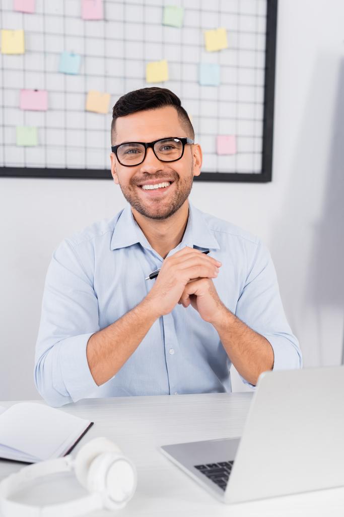 веселий бізнесмен в окулярах посміхається біля ноутбука на столі
 - Фото, зображення