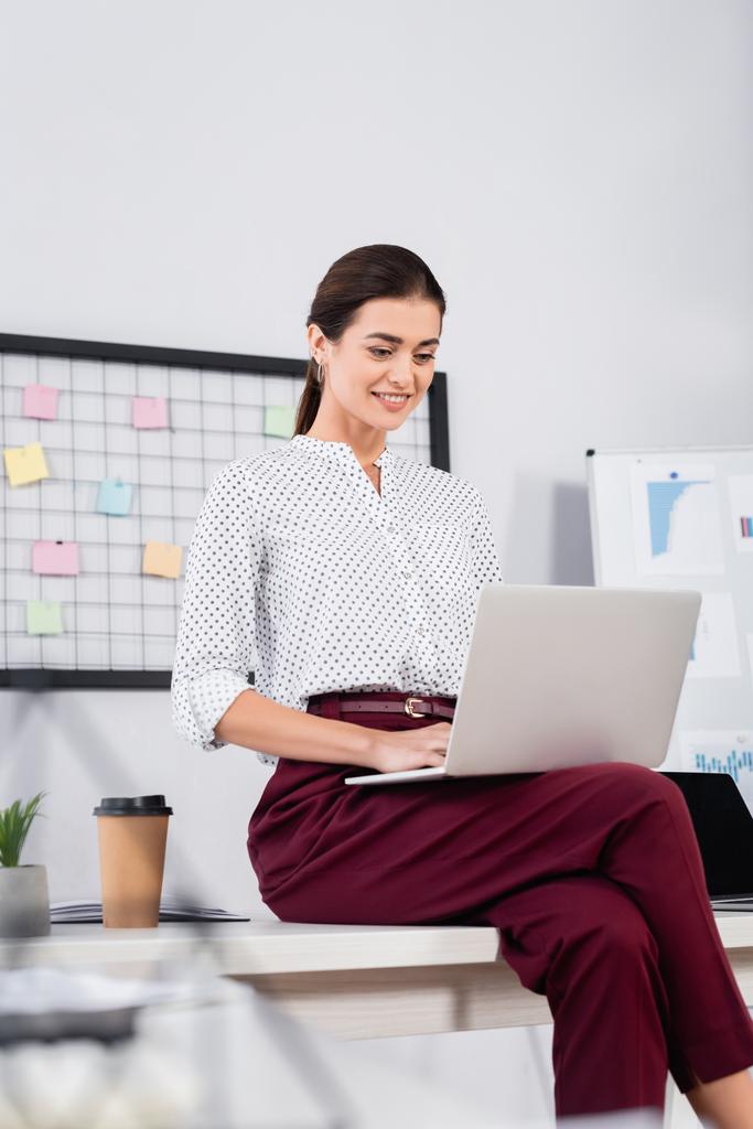 радостная деловая женщина смотрит на ноутбук и сидит на столе - Фото, изображение