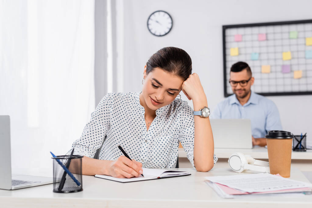 cheerful businesswoman writing in notebook near businessman on blurred background - Photo, Image