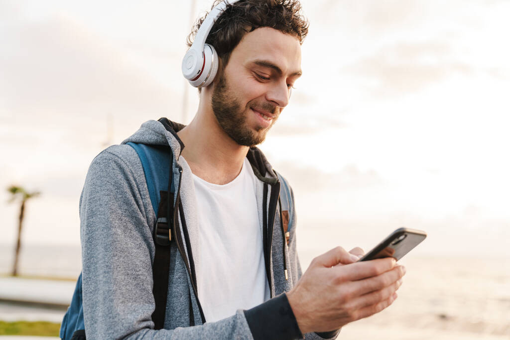 Усміхнений молодий хлопець в навушниках використовує мобільний телефон під час прогулянки на набережній
 - Фото, зображення