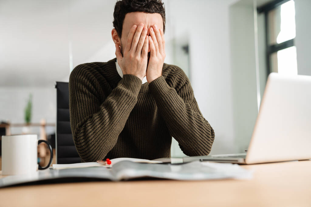 Hombre de negocios estresado mirando a la computadora portátil mientras está sentado en el escritorio de la oficina, cubriendo la cara con ojos - Foto, Imagen