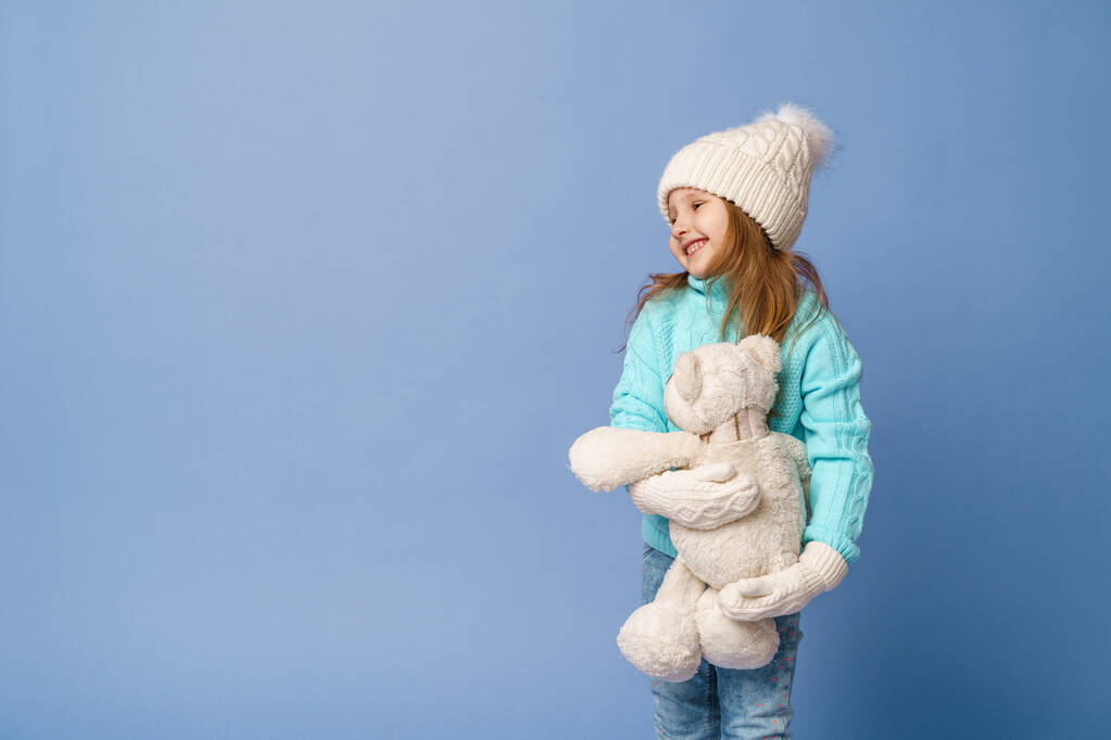 χαρούμενο 5χρονο κοριτσάκι με ξανθά μαλλιά σε πλεκτό πουλόβερ και γάντια, χαμογελάει και αγκαλιάζει ένα αρκουδάκι σε μπλε φόντο στο στούντιο. Το παιχνίδι είναι φίλος της. Χειμώνας. Σαββατοκύριακο διακοπών - Φωτογραφία, εικόνα