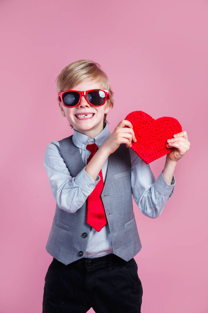 Valentines day. Stylish little boy in red sunglasses posing with heart on pink background - Photo, Image