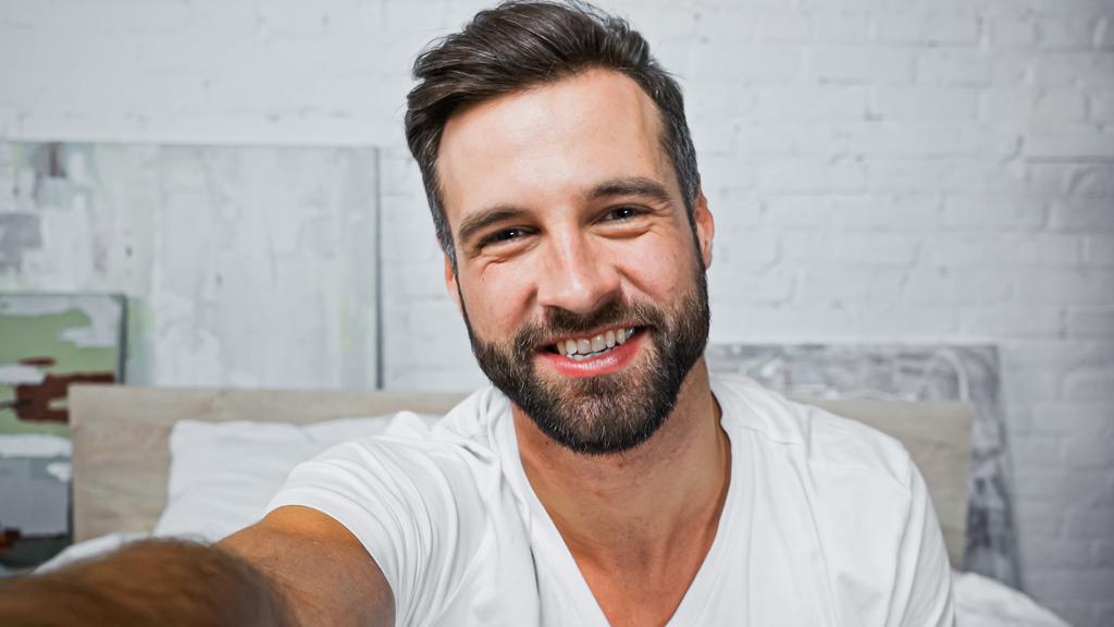幸せな髭を生やした男がベッドルームのカメラで笑顔で - 写真・画像
