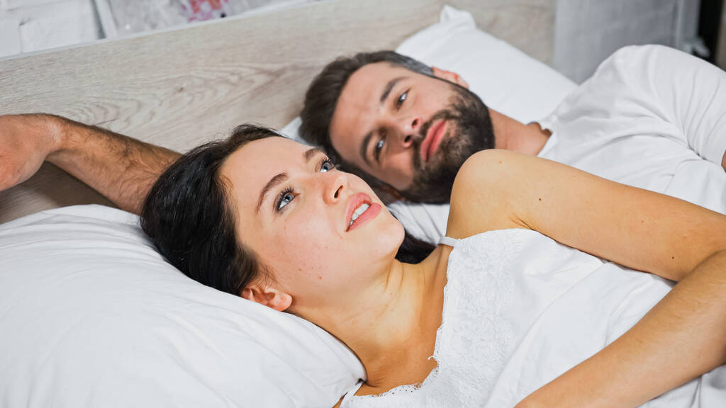 fiatal szakállas férfi néz barna nő fekszik a közelben az ágyban, homályos háttér - Fotó, kép
