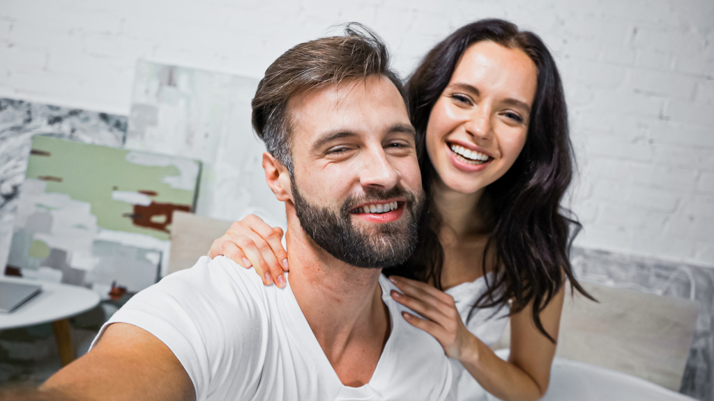 ευτυχισμένος γενειοφόρος άνδρας και μελαχρινή γυναίκα χαμογελά στην κάμερα στην κρεβατοκάμαρα - Φωτογραφία, εικόνα