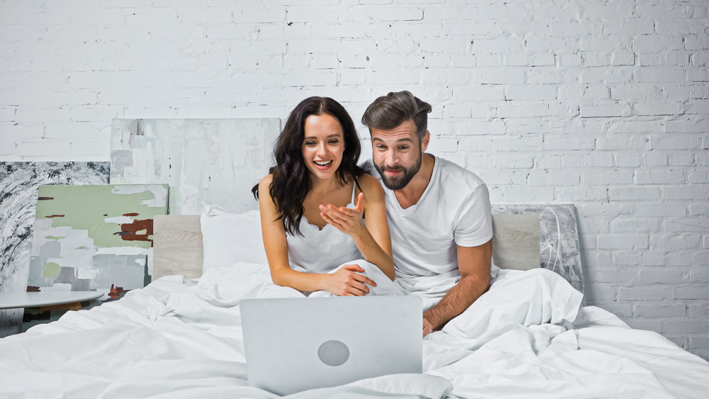 aufgeregte Frau zeigt mit der Hand auf Laptop, während sie sich Film mit Freund im Bett ansieht - Foto, Bild